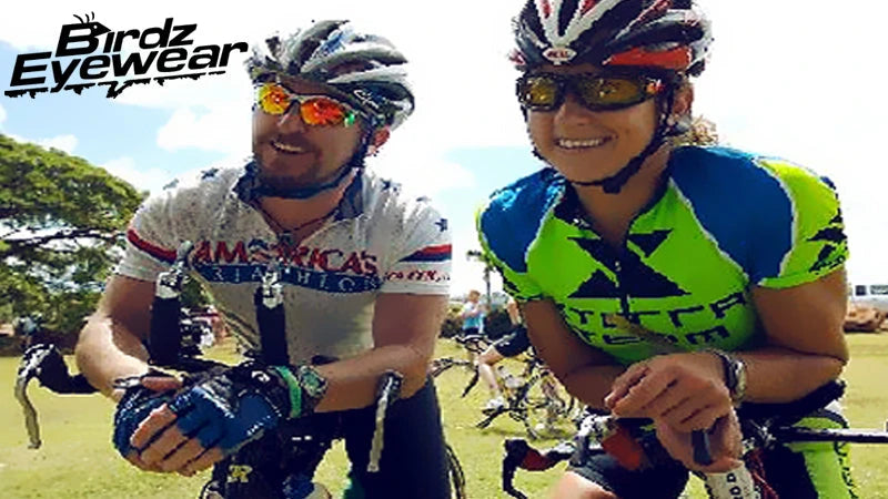 Two cyclists in colorful jerseys and helmets smile while leaning on their bikes in a sunny outdoor setting.