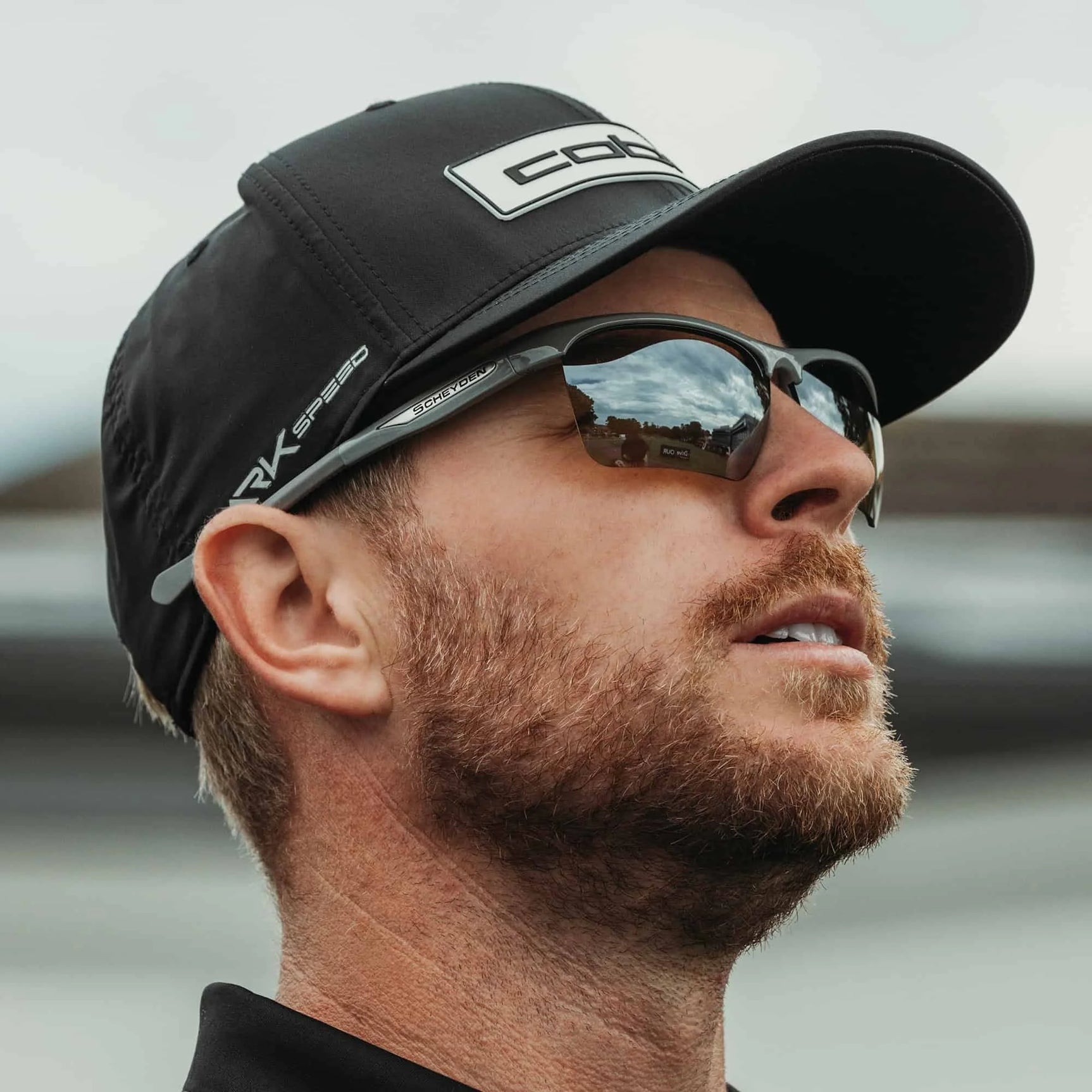 A bearded man, wearing a cap, gazes forward with the sky mirrored in his Scheyden CIA GRABBER Sunglasses.