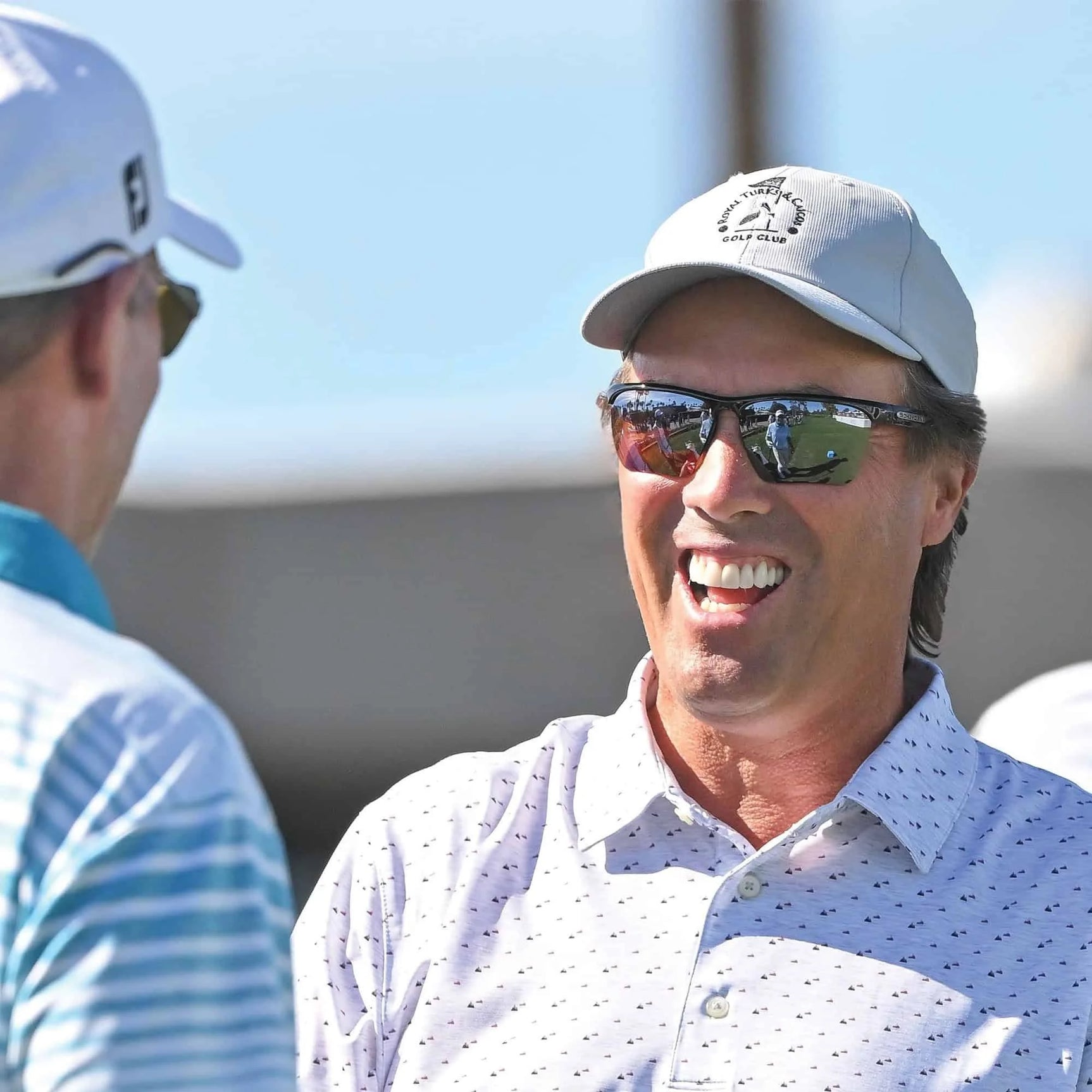 A man in a white golf shirt and cap, wearing Scheyden CIA GRABBER Sunglasses with TR90 composite frames, smiles while engaging in an outdoor conversation.