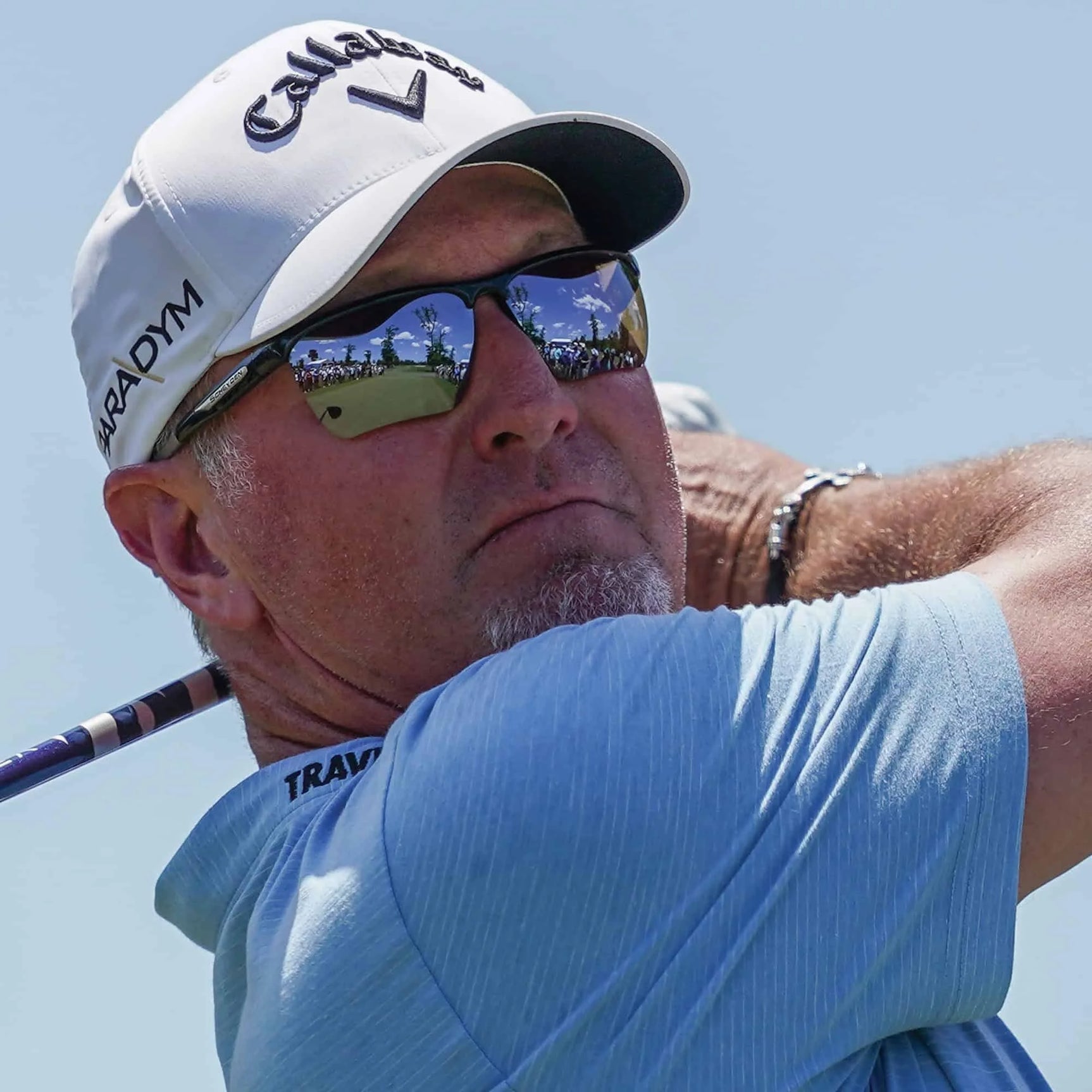 A person in Scheyden CIA GRABBER sunglasses with TR90 composite frames and a cap, holding a golf club mid-swing under the clear blue sky.